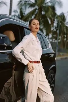 Женщина в белой одежде непринужденно опирается на дверцу черного автомобиля на фоне высоких пальм и солнечного неба, создавая стильную и расслабленную атмосферу