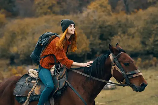 Рыжеволосая девушка в шапке и рюкзаке едет верхом на гнедом коне осенью