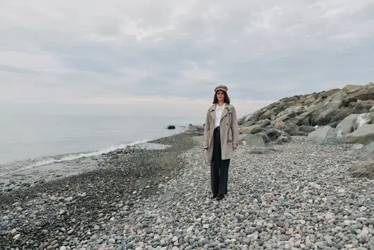 Молодая женщина в пальто и кепке стоит на каменистом берегу моря