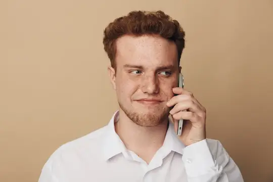 Рыжеволосый мужчина с веснушками в белой рубашке держит смартфон у уха, смотрит в сторону с легкой улыбкой
