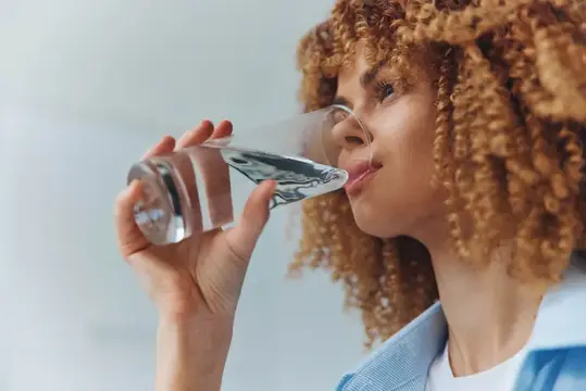 Кудрявая девушка пьёт воду из стеклянной бутылки