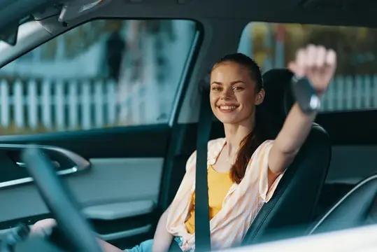 Улыбающаяся девушка за рулём автомобиля поднимает руку