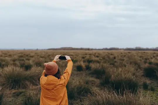 Женщина в жёлтом плаще фотографирует на телефон осеннее поле
