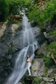 Девушка в белом стоит на камнях у подножия водопада