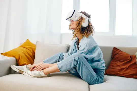 Улыбающаяся кудрявая девушка в VR-очках сидит на диване