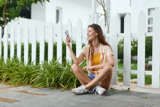 Улыбающаяся девушка сидит на дорожке у белого забора и смотрит в смартфон