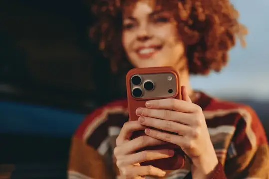 Женщина с вьющимися волосами улыбается, держа смартфон, момент селфи в женском лайфстайле, мягкая студийная атмосфера, теплый размытый фон, повседневный портрет дома, нежное выражение лица, крупный…