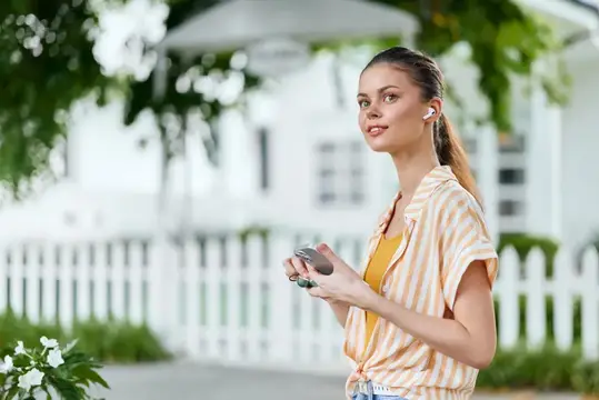 Молодая женщина в наушниках со смартфоном на фоне белого забора