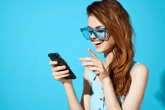 Рыжеволосая девушка в очках улыбается, глядя в смартфон на голубом фоне