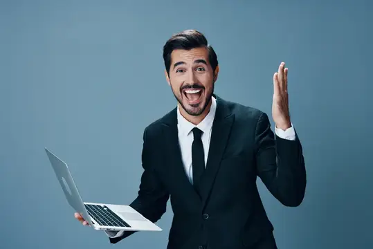 Мужчина в деловом костюме смотрит на ноутбук и работает онлайн через интернет, совершает видеозвонок, счастливо улыбается, показывая зубы, поднимает руки вверх от радости на синем фоне