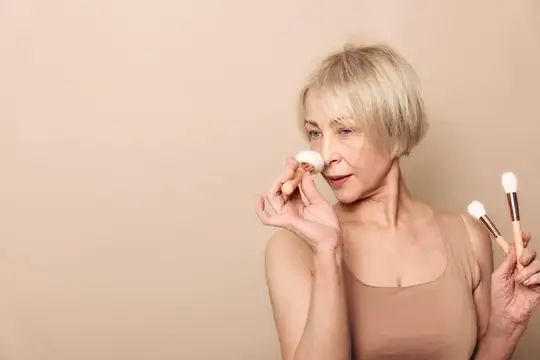 Светловолосая пожилая женщина, держа кисти для макияжа, аккуратно наносит пудру на лицо