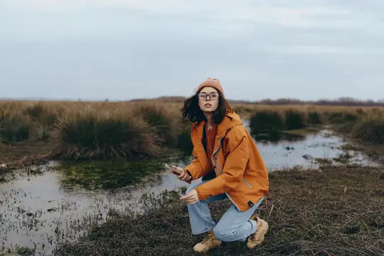Женщина на открытом воздухе сидит на корточках у речной воды в оранжевой куртке на фоне облачного осеннего пейзажа