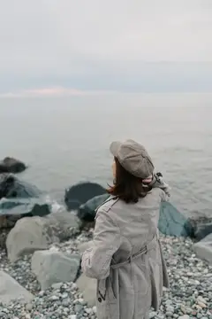 Вид сзади на женщину в сером пальто и клетчатой шляпе, стоящую на каменистом берегу и смотрящую на спокойное море под облачным небом