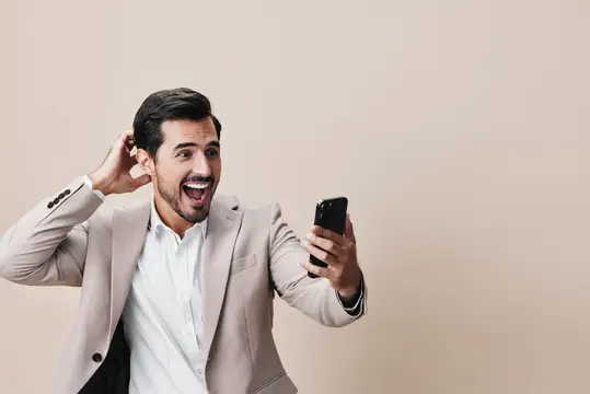 Эмоциональный мужчина в бежевом пиджаке смотрит в смартфон и жестикулирует