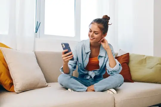 Улыбающаяся девушка со смартфоном сидит по-турецки на диване