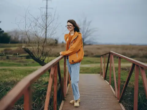 Улыбающаяся девушка в очках и оранжевой куртке стоит на деревянном мостике