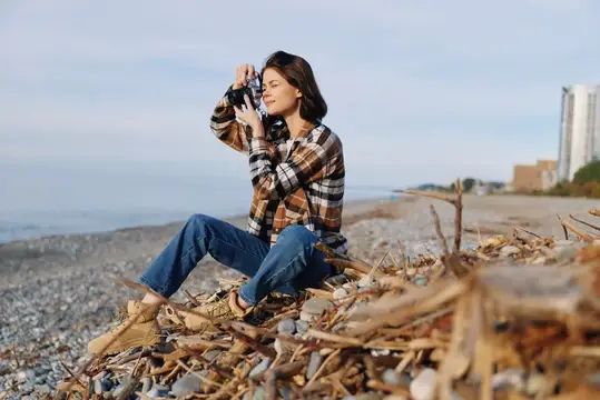 Молодая женщина с фотокамерой сидит на камнях у моря