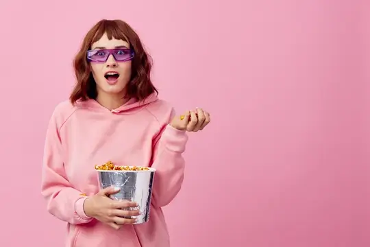 Удивлённая девушка в 3D-очках с ведром попкорна на розовом фоне