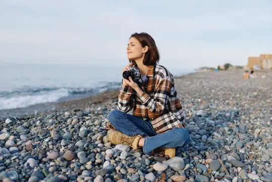 Молодая женщина с фотокамерой сидит на галечном пляже у моря