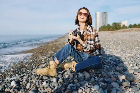 Улыбающаяся женщина с фотокамерой сидит на каменистом берегу