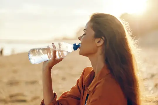 Молодая женщина пьёт воду из бутылки на закатном пляже