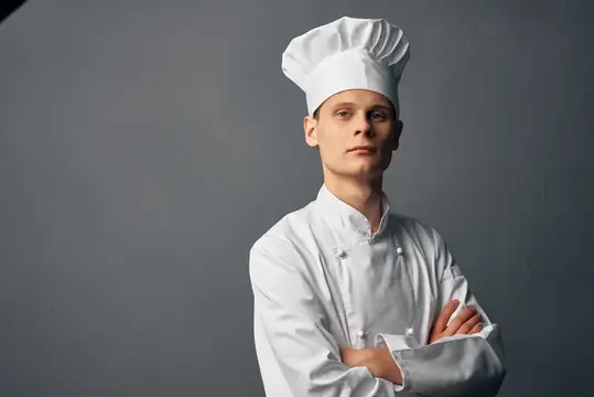 Молодой повар в белом колпаке со скрещёнными руками на сером фоне