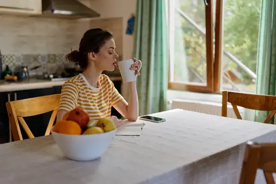 Молодая женщина пьёт кофе за столом с фруктами у окна