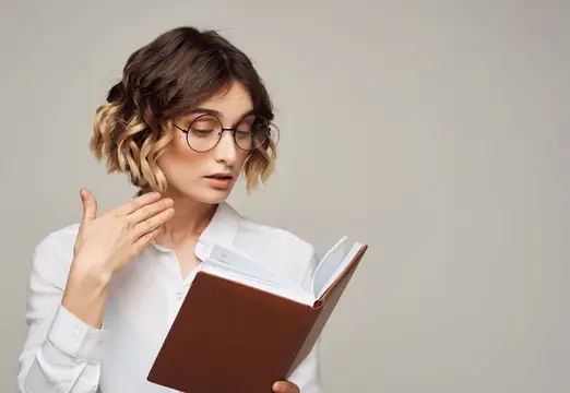 Молодая женщина в очках увлечённо читает книгу на светлом фоне