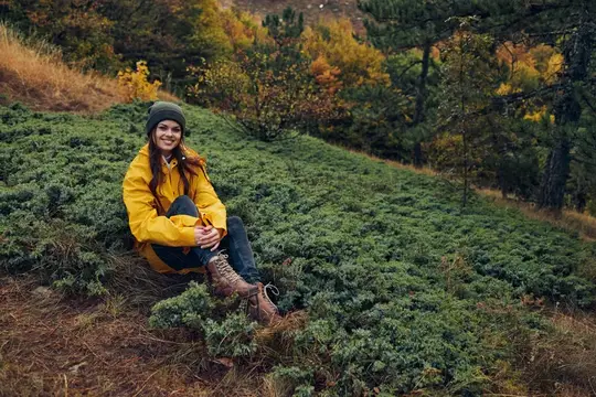 Молодая женщина в жёлтой куртке сидит на холме в лесу, наслаждаясь спокойным моментом среди красоты природы