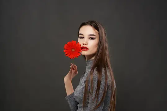Симпатичная брюнетка с красным цветком у лица на тёмном фоне