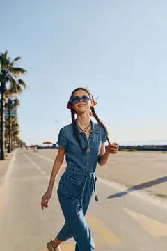 Улыбающаяся девушка с косичками и в солнцезащитных очках идёт по асфальту вдоль пальм
