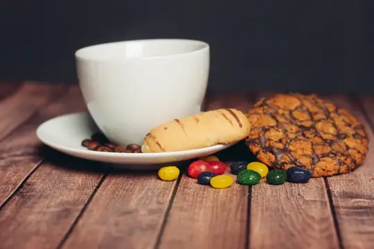 Белая чашка на блюдце с печеньем и разноцветными конфетами на деревянном столе