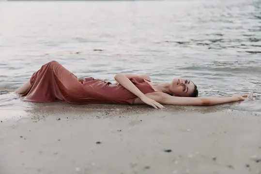 Девушка в шелковом платье лежит на мокром песке у воды