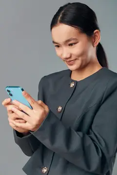 Деловая женщина в темном пальто сосредоточена на экране смартфона, держа устройство обеими руками на однотонном фоне