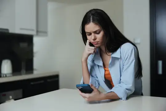 Задумчивая темноволосая женщина со смартфоном за столом на кухне