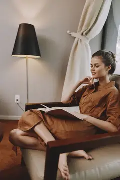 Молодая женщина в коричневом платье читает книгу в кресле у торшера