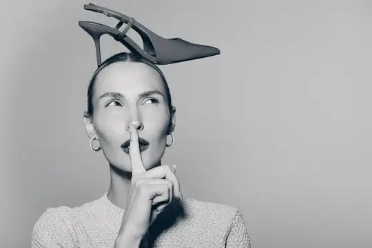 Портрет женщины-европеоида со светлой кожей и каштановыми волосами, делающей жест молчания пальцем и балансирующей чёрной туфлей на каблуке на голове