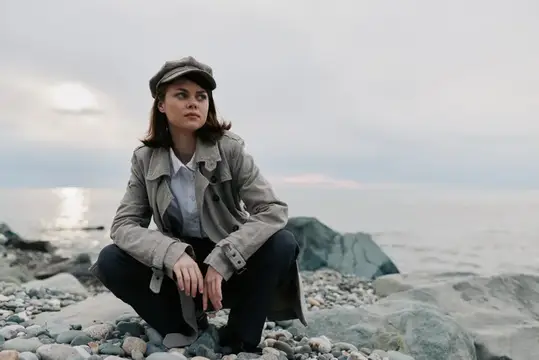 Молодая женщина в пальто и кепке сидит на корточках на каменистом берегу у спокойного моря на облачном закате