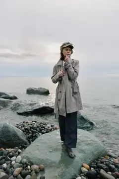 Девушка в плаще и кепке стоит на камне у моря