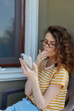 Кудрявая девушка в очках смотрит в смартфон у окна