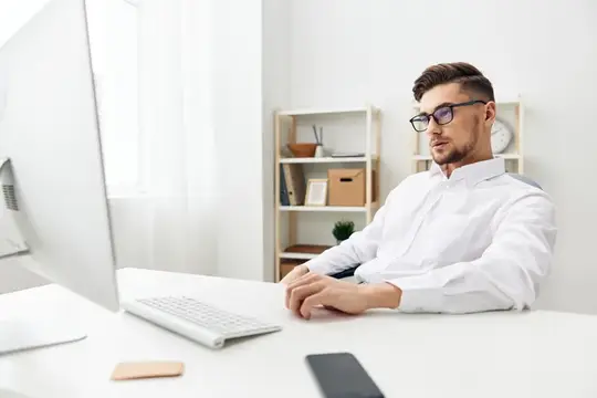 Молодой мужчина в очках и белой рубашке работает за компьютером в светлом офисе