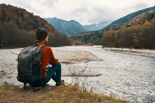Женщина с рюкзаком, в куртке и шапке стоит на берегу реки в горах, вид сбоку