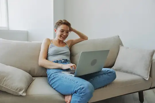 Улыбающаяся девушка в очках работает за ноутбуком на диване
