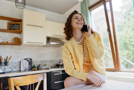 Улыбающаяся кудрявая девушка в жёлтом кардигане говорит по телефону на светлой кухне