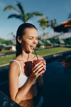 Улыбающаяся женщина у бассейна потягивает смузи, теплый солнечный свет, прозрачная вода, тропическая атмосфера, непринужденное отпускное настроение, демонстрирующее яркий образ жизни и отдых на…