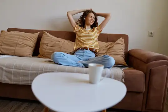 Улыбающаяся кудрявая девушка отдыхает на диване с чашкой кофе