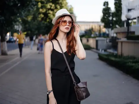 Рыжеволосая девушка в шляпе и очках поправляет причёску на городской улице