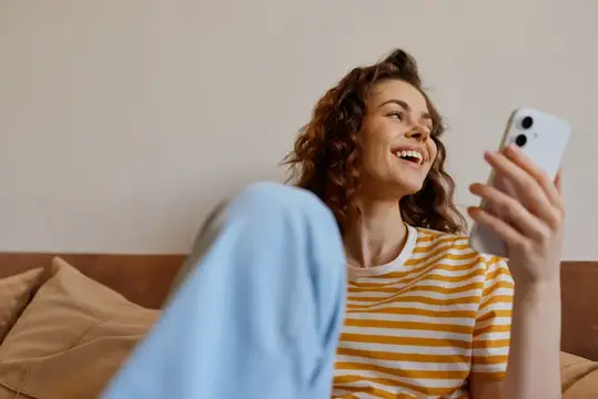 Кудрявая девушка смеётся, глядя в смартфон на диване