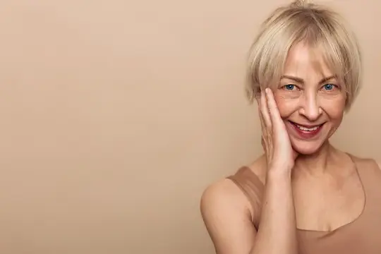 Улыбающаяся блондинка в возрасте с рукой у щеки на бежевом фоне
