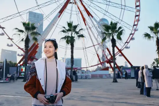 Улыбающаяся азиатская девушка с фотокамерой на фоне колеса обозрения